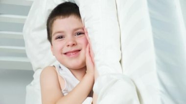 Cute 5 year old boy lies on his side on a bed on a pillow and smiles looking into the camera.