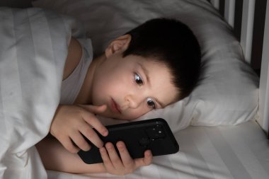 A 5 year old boy plays games on his phone while lying in bed. Gadgets in bed before bedtime in a child.
