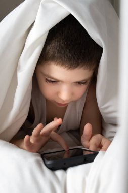 A 5 year old boy plays games on his phone while lying in bed. Gadgets in bed before bedtime in a child.