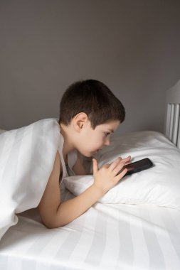 A 5 year old boy plays games on his phone while lying in bed. Gadgets in bed before bedtime in a child.