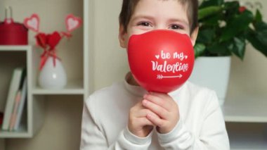 Cute 5 year old boy holds a balloon with the text be my valentine and smiles looking into the camera.