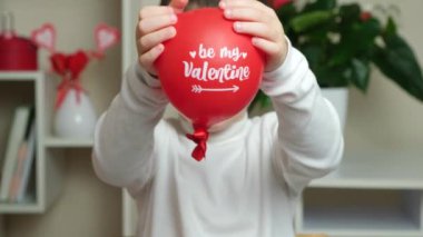 Cute 5 year old boy holds a balloon with the text be my valentine and smiles.