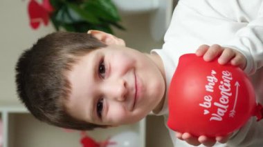 Cute 5 year old boy holds a balloon with the text be my valentine and smiles looking into the camera.