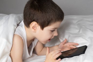 A 5 year old boy plays games on his phone while lying in bed. Gadgets in bed before bedtime in a child.