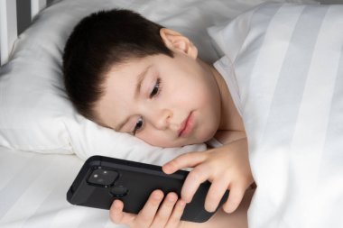 A 5 year old boy plays games on his phone while lying in bed. Gadgets in bed before bedtime in a child.