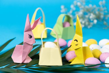 Two figures of Easter bunnies made of paper, origami crafts and a basket with eggs on a blue background.