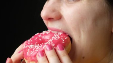 A woman eats a delicious red doughnut, bites and chews it appetizingly, close-up. High-calorie sweet food, diabetes.