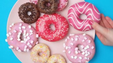 Bir tabakta kırmızı, pembe ve çikolata kaplı çörekler, bir kadın bir çörek alır, mavi arka plan, üst manzara.