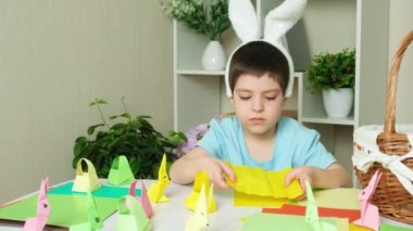 Tavşan kulaklı anaokulu çocuğu Paskalya zanaatı, origami tavşan figürleri ve kağıt sepetleri yapıyor..