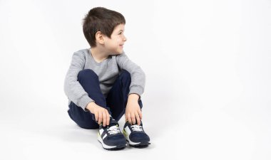 Gri eşofmanlı, mavi eşofmanlı ve beyaz arka planda spor ayakkabılı küçük bir çocuk oturur ve yüzünü çevirir..