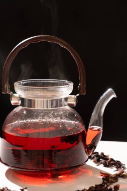 Red hibiscus tea from the petals of a Sudanese rose in a glass teapot, steam from a teapot on a black background.