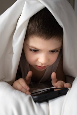 A 5 year old boy plays games on his phone while lying in bed. Gadgets in bed before bedtime in a child.