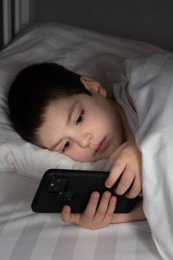 A 5 year old boy plays games on his phone while lying in bed. Gadgets in bed before bedtime in a child.