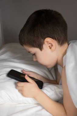 A 5 year old boy plays games on his phone while lying in bed. Gadgets in bed before bedtime in a child.