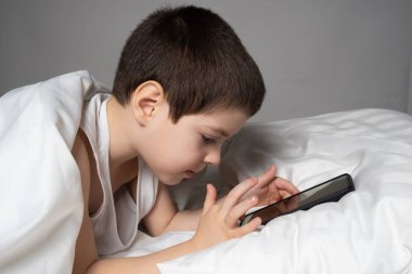 A 5 year old boy plays games on his phone while lying in bed. Gadgets in bed before bedtime in a child.