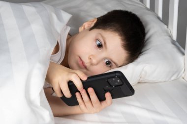 A 5 year old boy plays games on his phone while lying in bed. Gadgets in bed before bedtime in a child.