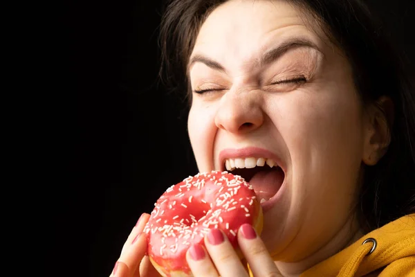 A woman bites a large red donut, a black background, a place for text. Gluttony, overeating and sugar addict