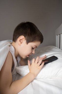 A 5 year old boy plays games on his phone while lying in bed. Gadgets in bed before bedtime in a child.