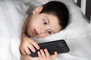 A 5 year old boy plays games on his phone while lying in bed. Gadgets in bed before bedtime in a child.