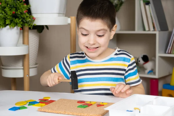 A preschool child plays with a mosaic, builds figures on a board, hammering nails with a hammer on multi-colored parts of the designer.