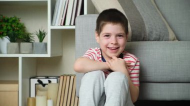 5 yaşında bir çocuk gülüyor, elleriyle yüzünü örtüyor, ve evde kanepede otururken kameraya bakıp gülümsüyor..