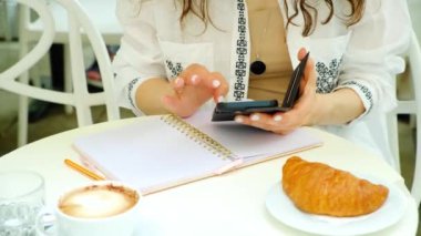 Açık hava kafede oturan bir kadın telefon, defter, kahve ve kruvasan kullanıyor..