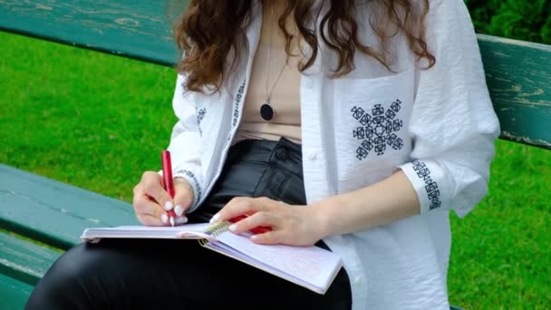 一位年轻女子坐在公园的长椅上 一边用笔记本里的钢笔写字 — 图库视频影像