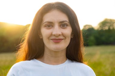 35 yaşında esmer bir kadının portresi. Doğaya gülümseyen kameraya bakıyor.