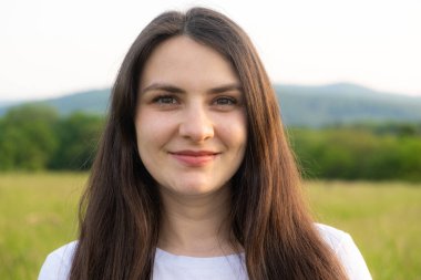 30 yaşında esmer bir kadının portresi. Doğada gülümseyen kameraya bakıyor.