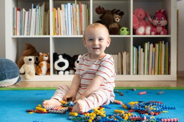 Bir yaşındaki bir bebek halının üzerinde oturur ve evde tasarımcıyla oynar.