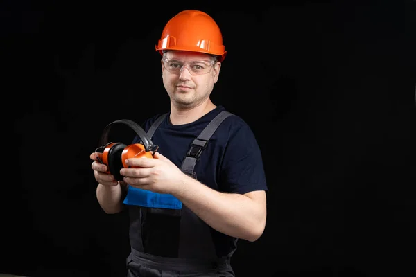 Builder in a protective helmet with noise-cancelling headphones in his hands on a black background, copy space for text.