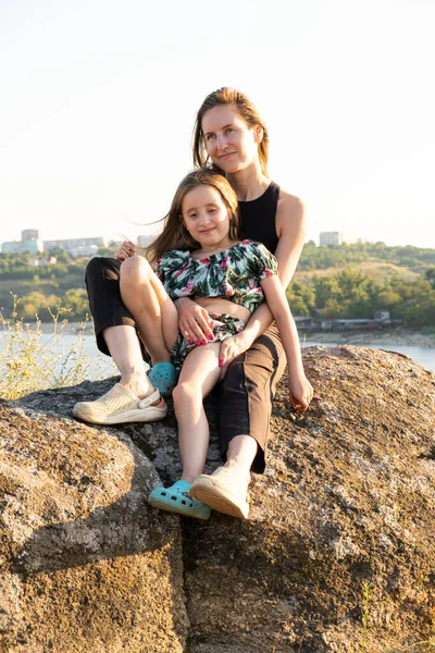 Happy mom and daughter relaxing on rocks in nature against the backdrop of the river. Happy Motherhood, Mother's Day.