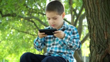 Yazın parkta otururken akıllı telefondan oyun oynayan küçük bir çocuk..