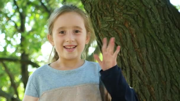 Маленькая Девочка Машет Рукой Глядя Камеру Сидя Возле Дерева Парке — стоковое видео