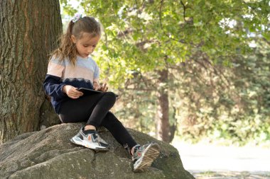 Küçük kız yazın parkta otururken bir tablet kullanır.