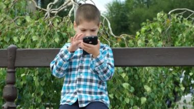 Küçük bir çocuk yazın parkta bir bankta otururken akıllı telefon kullanır.