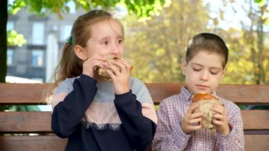 Küçük çocuklar, bir erkek ve bir kız hamburger yer, yazın bir bankta oturur.