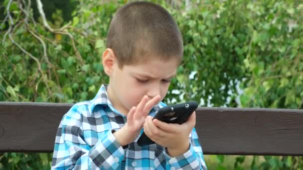 小さな男の子は夏に公園のベンチに座っている間スマートフォンを使用します — ストック動画