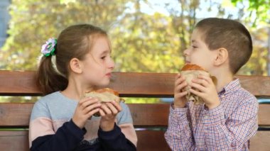 Küçük çocuklar, bir erkek ve bir kız hamburger yer, yazın bir bankta oturur.