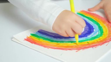 Küçük bir çocuk masada otururken çizim defterine keçeli kalemlerle gökkuşağı çiziyor..