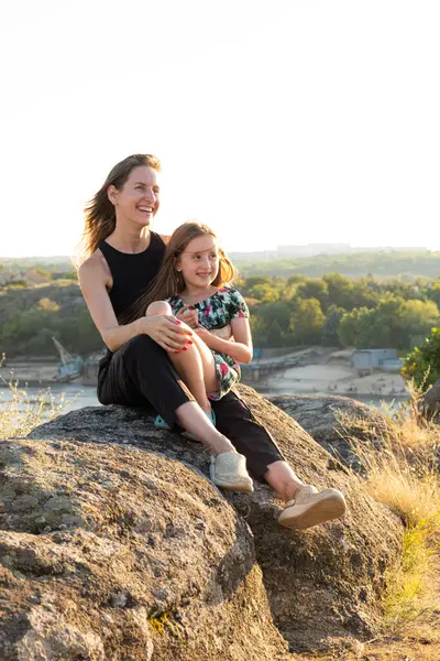 Happy mom and daughter relaxing on rocks in nature against the backdrop of the river. Happy Motherhood, Mothers Day.