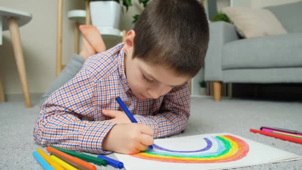 소년은 집에서 카펫에 누워있는 스케치북에 펜으로 무지개를 그립니다 — 비디오