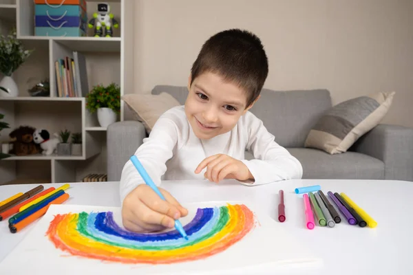 A small child draws a rainbow with felt-tip pens in a sketchbook while sitting at a table.