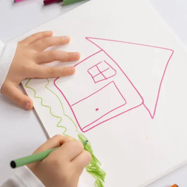 Little child drawing with felt-tip pens a house with grass in a sketchbook