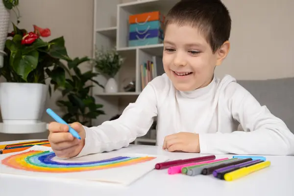 A small child draws a rainbow with felt-tip pens in a sketchbook while sitting at a table.