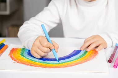 Küçük bir çocuk masada otururken çizim defterine keçeli kalemlerle gökkuşağı çiziyor..
