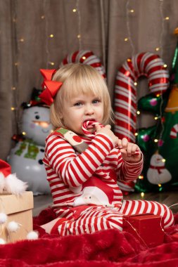 Noel 'de evde hediyelerin arasında oturmuş lolipop yiyen iki yaşında bir kız..