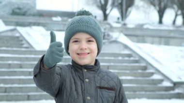 Kış parkında merdivenlerin yanında elleri süper olan altı yaşında bir çocuk baş parmak işareti yapıyor.
