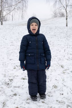 Sıcak tulumlu, dilini çıkarıp kış parkında somurtan komik küçük çocuk.