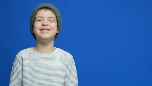 帽子と手袋のかわいい子供は 青い背景に彼の手で笑って彼の顔を覆う — ストック動画